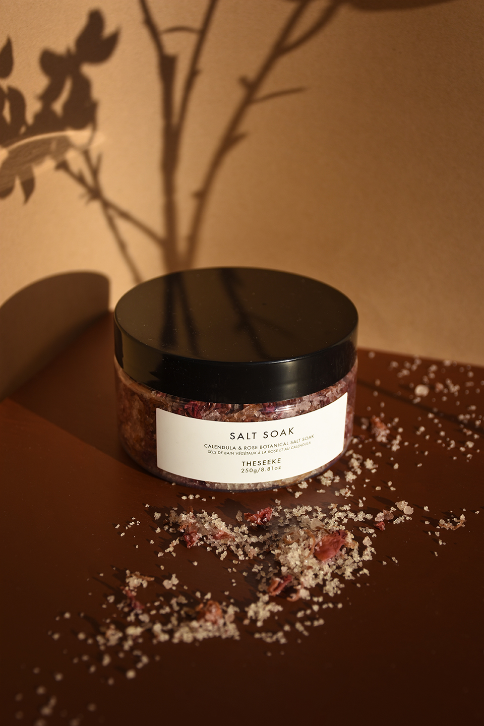 A container of bath salts with some of the salts spread across the foreground and a shadow of flowers in the background